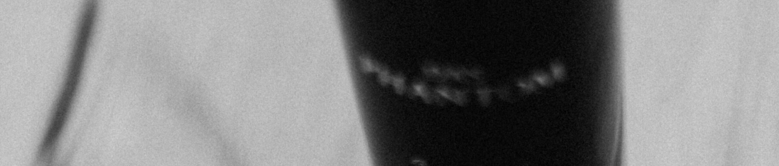 Unscharfes Schwarz-Weiß-Bild, das ein dunkles Objekt mit schwachem Text, möglicherweise ein Mikrofon, vor einem hellgrauen Hintergrund zeigt. Abstrakter und geheimnisvoller Ton.