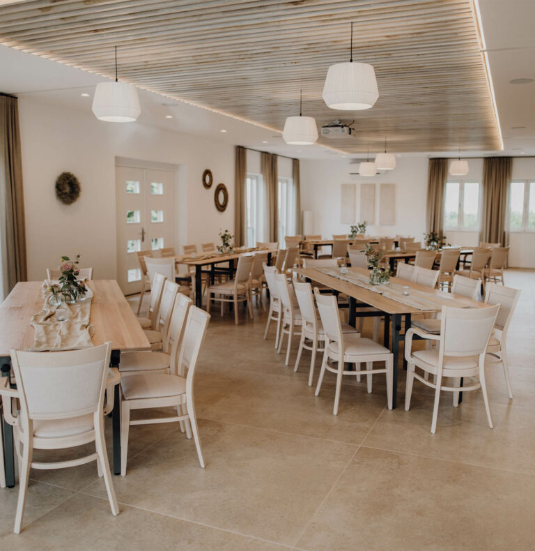 Ein geräumiges, elegantes Esszimmer mit Holztischen und beigen Stühlen. Sanfte Beleuchtung, dekorative Kränze und neutrale Töne schaffen ein ruhiges, einladendes Ambiente.