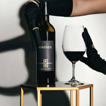 Die behandschuhten Hände halten elegant eine Weinflasche mit der Aufschrift „Forever“ und ein gefülltes Glas auf einem schlichten Goldtisch. Die Szene strahlt Raffinesse und Luxus aus.