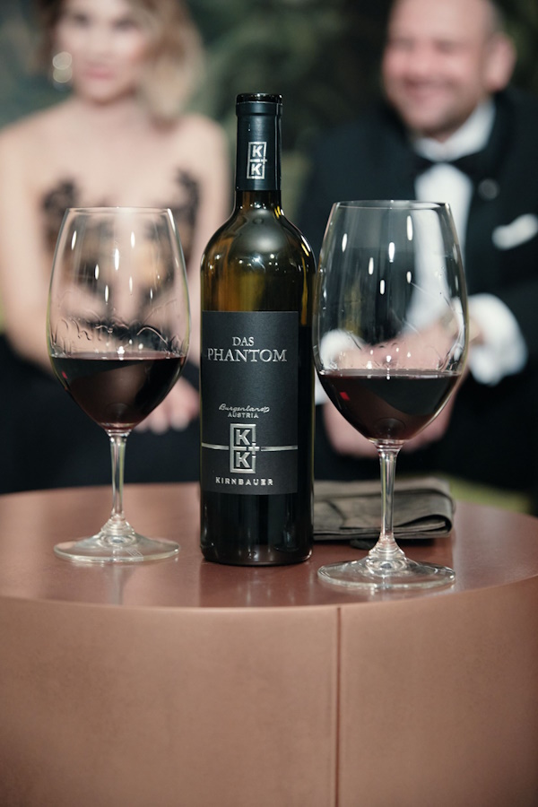 Eine Flasche des Rotweins „Das Phantom“ von Kirnbauer steht flankiert von zwei Gläsern auf einem Tisch. Im unscharfen Hintergrund erscheinen ein Mann und eine Frau, die förmlich gekleidet sind.