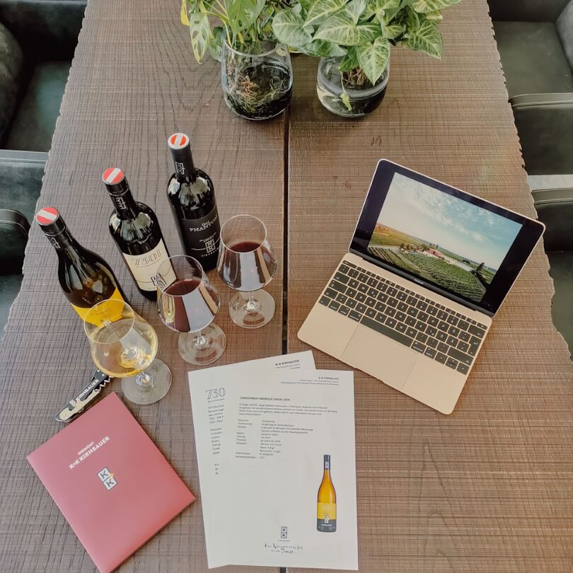 Drei Weinflaschen und -gläser, ein Laptop, auf dem ein Weinberg abgebildet ist, Papiere, ein Korkenzieher und ein rotes Büchlein auf einem Holztisch, daneben eine Pflanze.