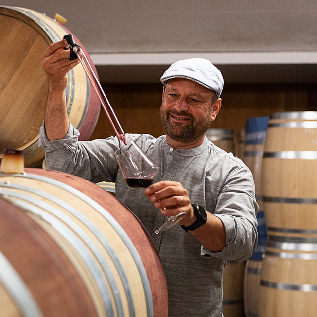 Der Winzer Markus Kirnbauer probiert Rotwein mit einer Pipette aus einem Fass in einer Weinkellerei. Er hält ein Glas in der Hand, umgeben von Holzfässern, die eine handwerkliche Atmosphäre vermitteln.